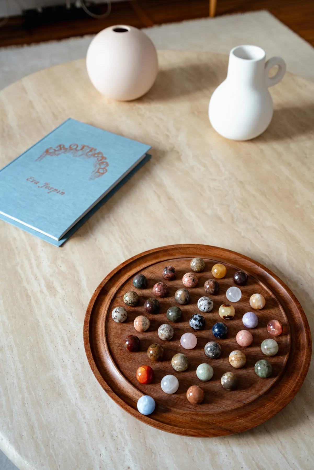 Una mesa muestra un libro, dos jarrones blancos y una bandeja redonda con piedras de colores.
