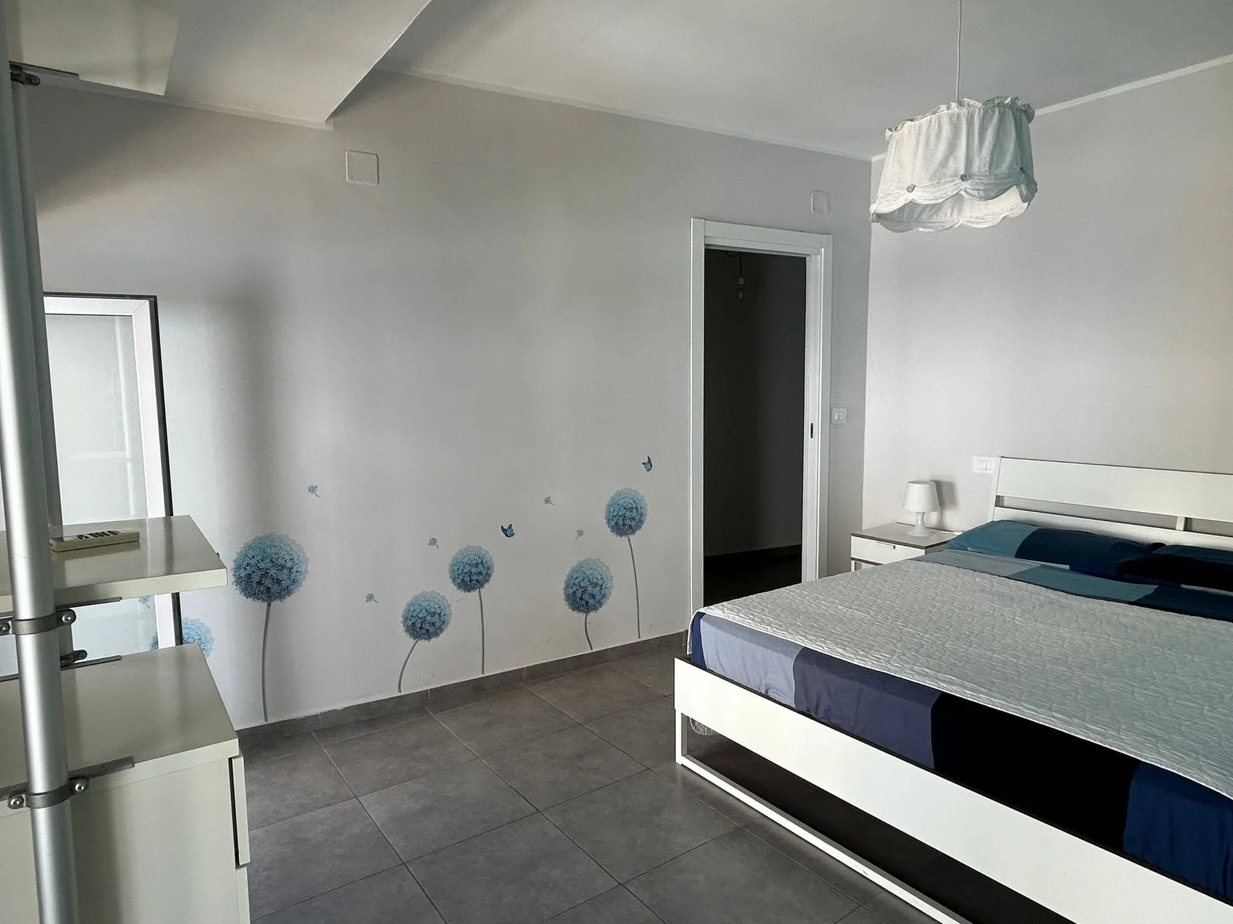 Un dormitorio con una cama, suelo de baldosas grises y pequeñas flores azules decorativas en la pared.