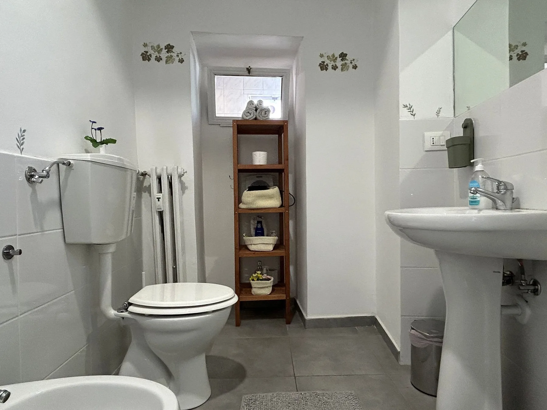 Un baño alicatado con inodoro, lavabo y estantería de madera bajo una ventana pequeña.