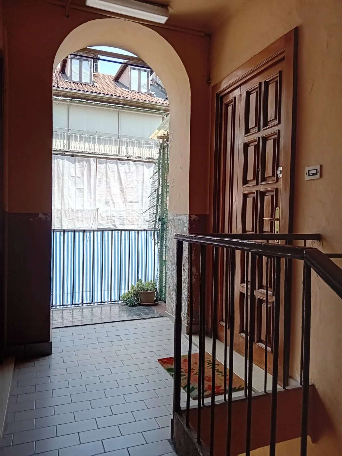 Un pasillo interior da a un patio luminoso, con suelo de baldosas y una barandilla oscura.