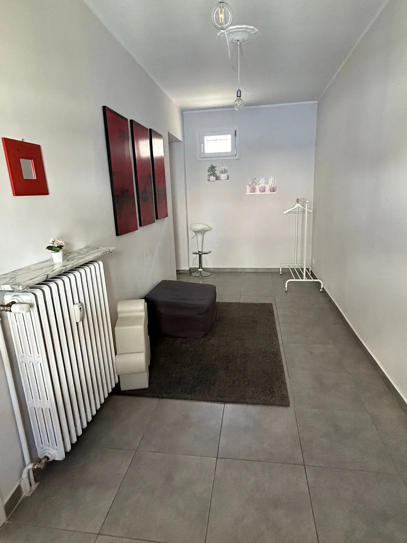 Una habitación interior estrecha con un radiador, una alfombra oscura y un taburete pequeño.