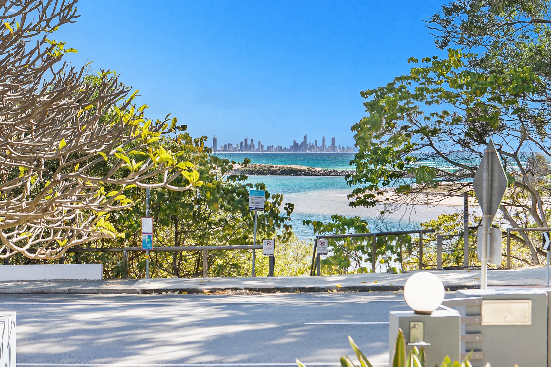 A coastal viewpoint overlooks turquoise water, a rocky shoreline, and a distant city skyline.