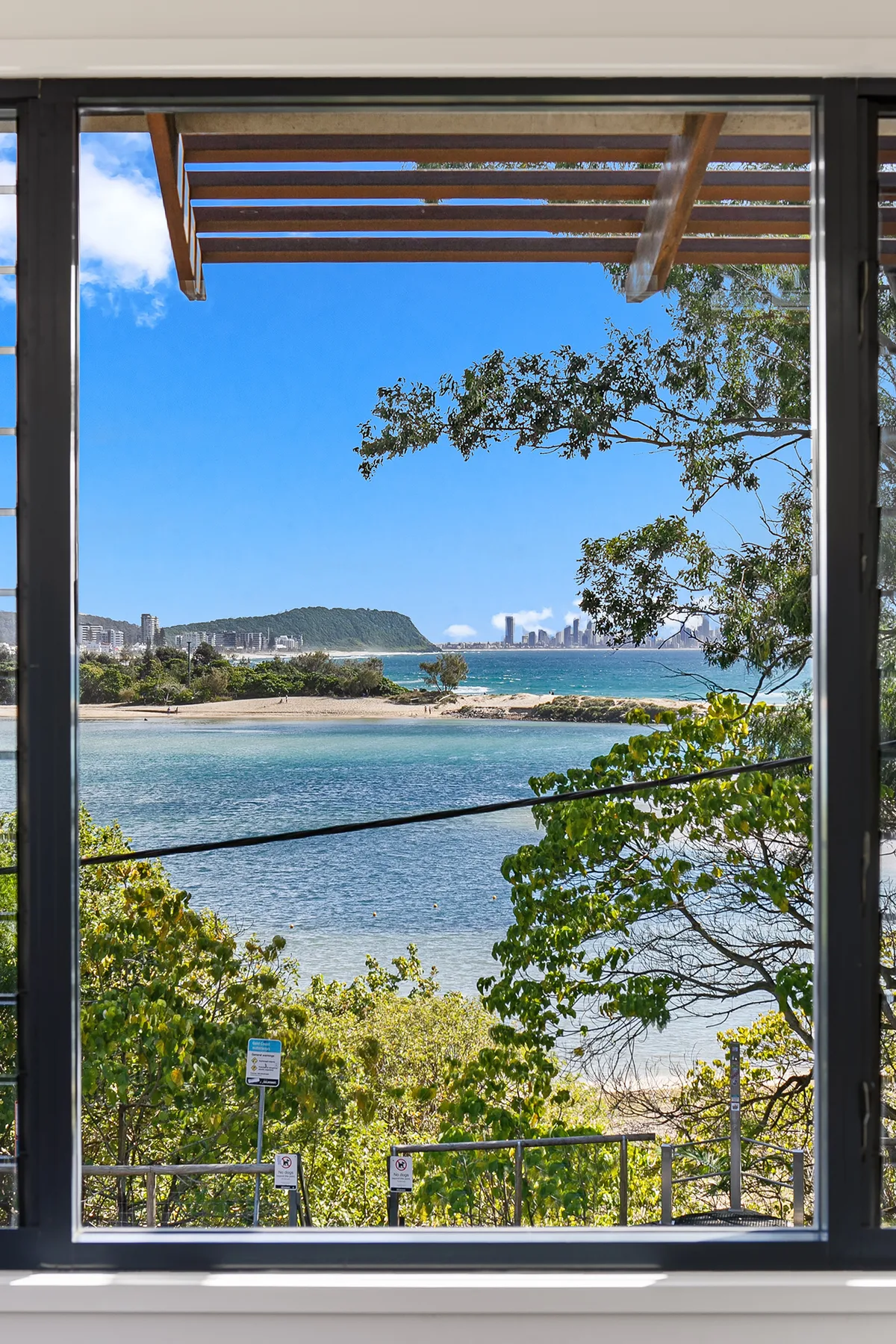 A large window overlooks a blue bay, with trees and a pergola visible outside.
