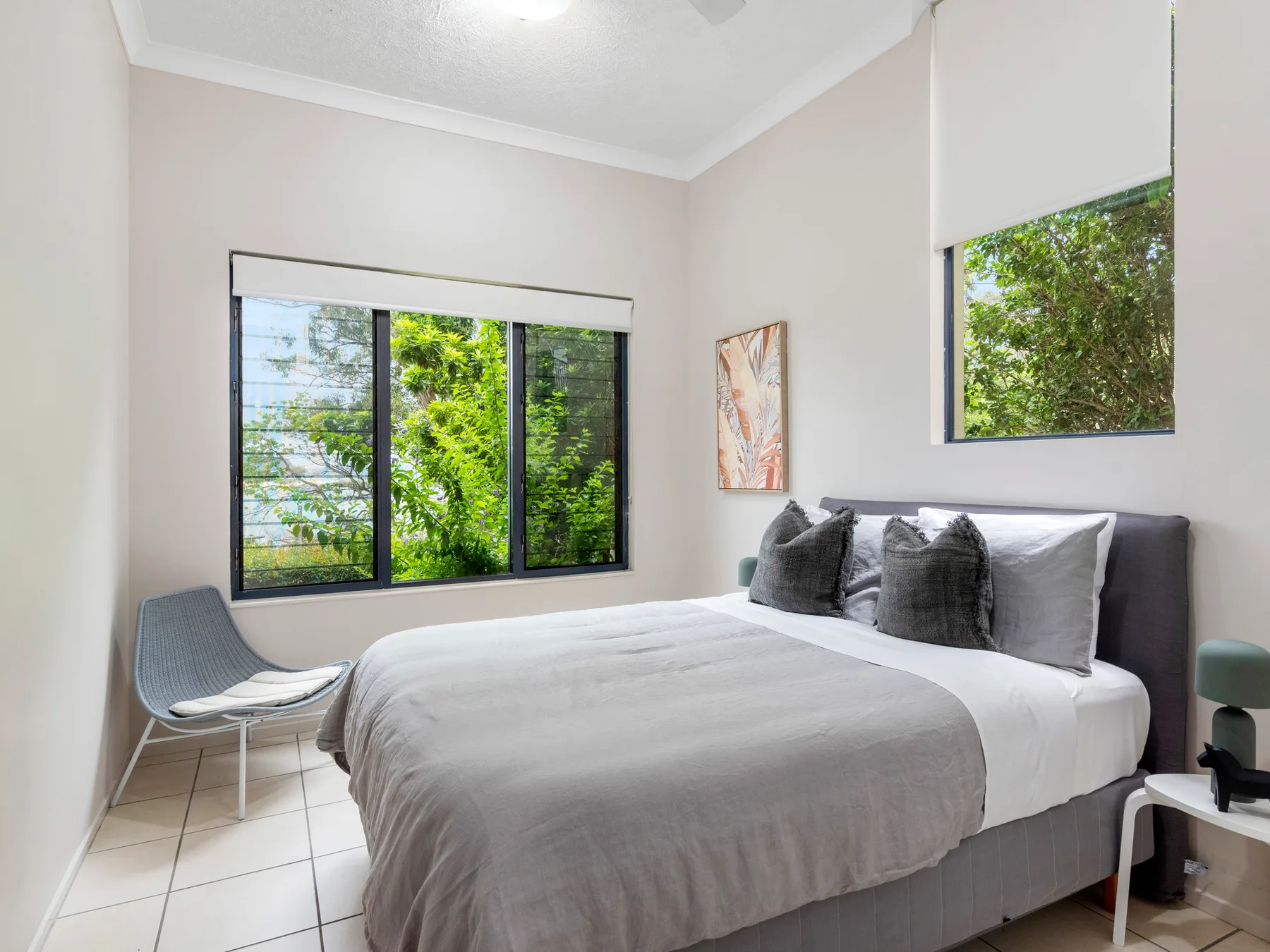 A bright bedroom with a large bed, gray bedding, and windows overlooking greenery.