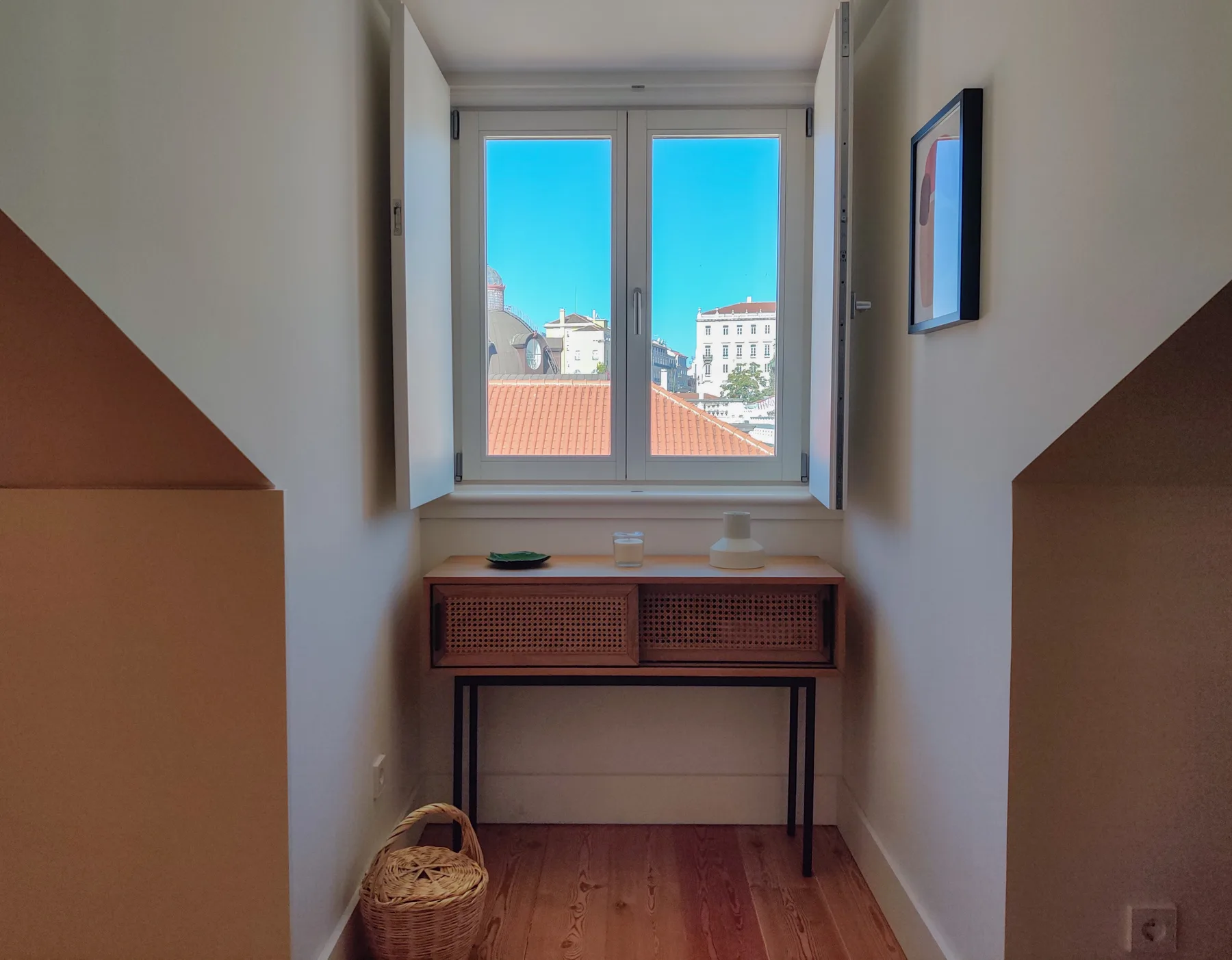 A narrow room with a desk beneath a window overlooking rooftops.