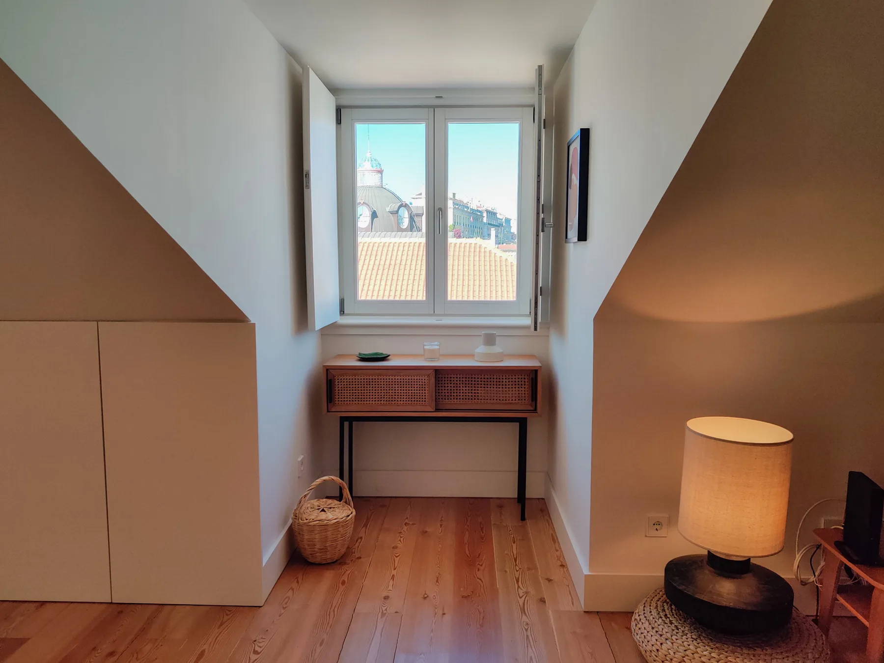 A small bedroom nook with a window above a narrow desk and warm wood flooring.