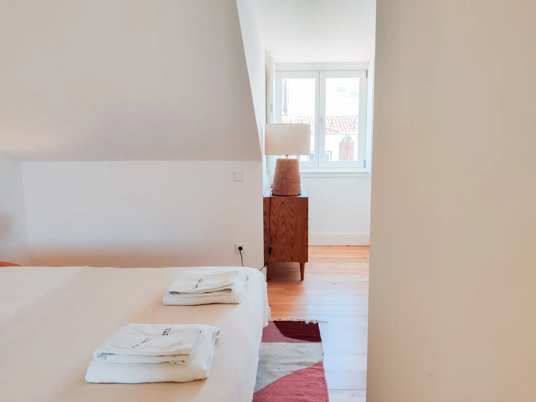 A bedroom with a bed, folded towels, and a wooden bedside table near a window.