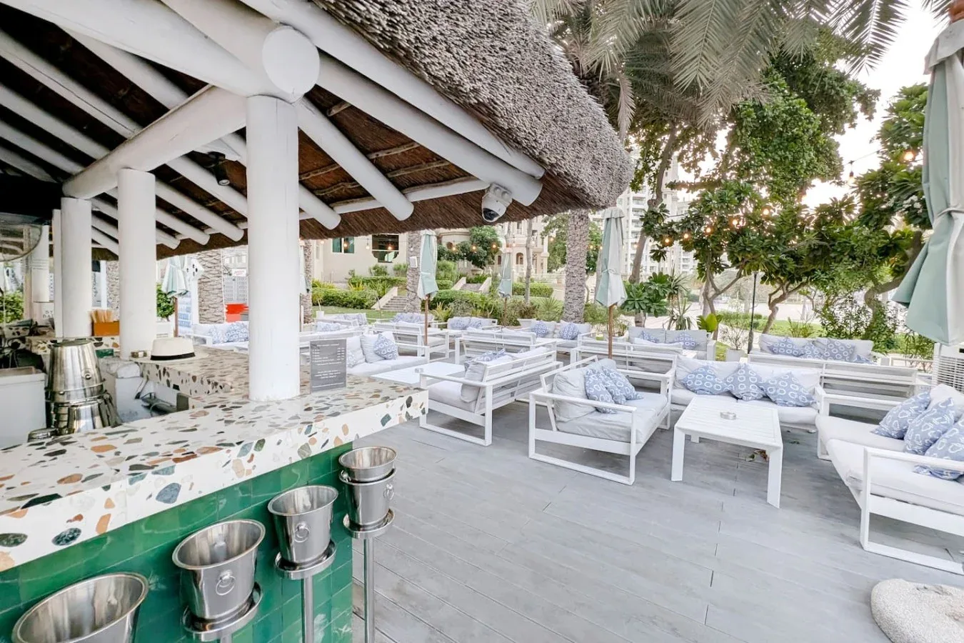 An outdoor lounge area has white seating beneath a thatched canopy.