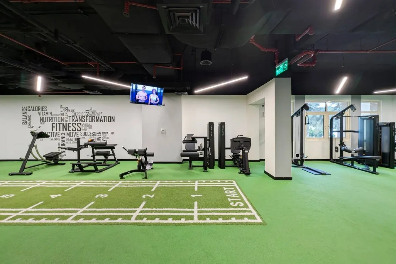 A gym with exercise machines on green flooring and a marked workout area.
