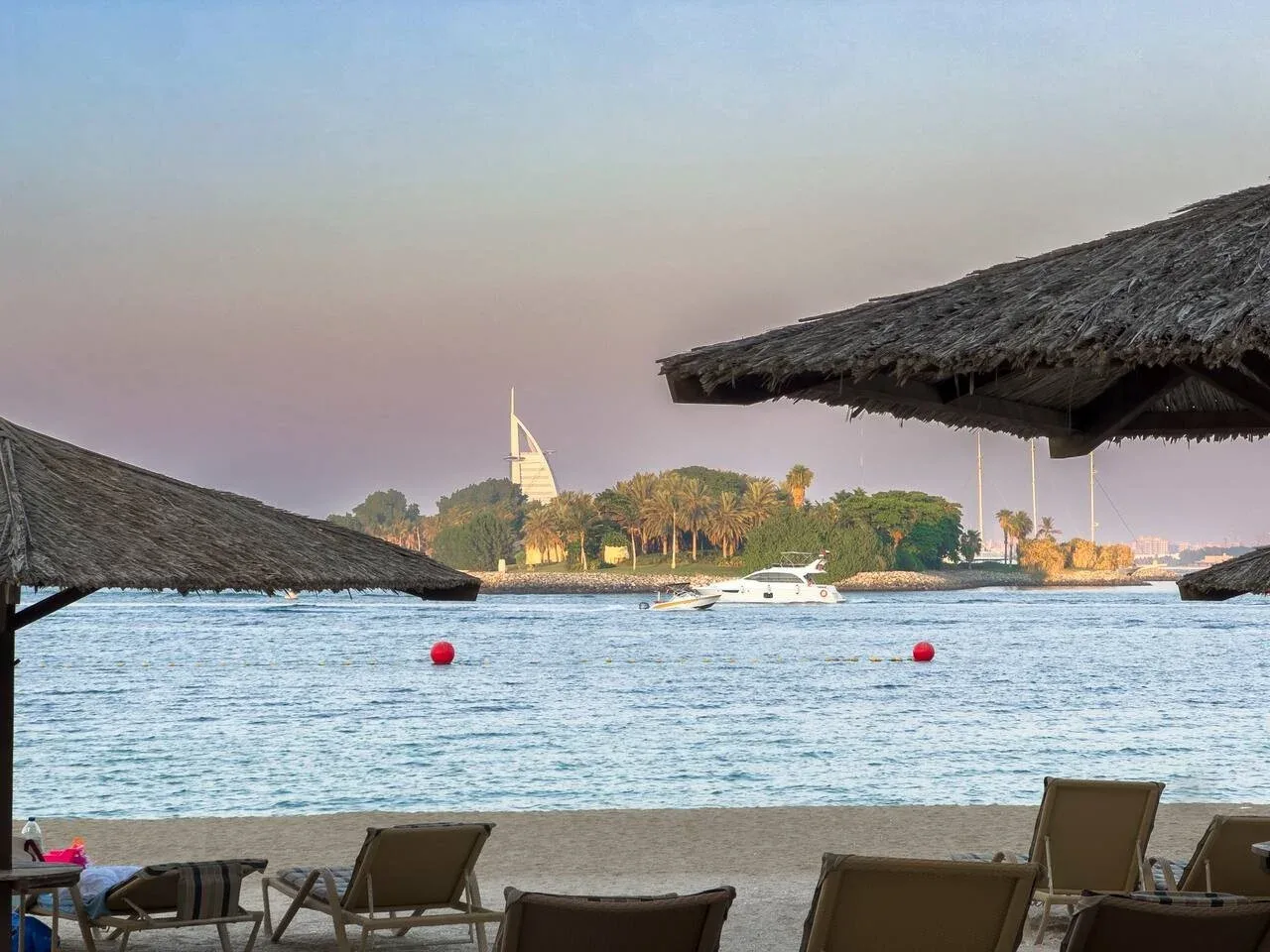A waterfront beach with lounge seating beneath thatched shade and boats on the water.