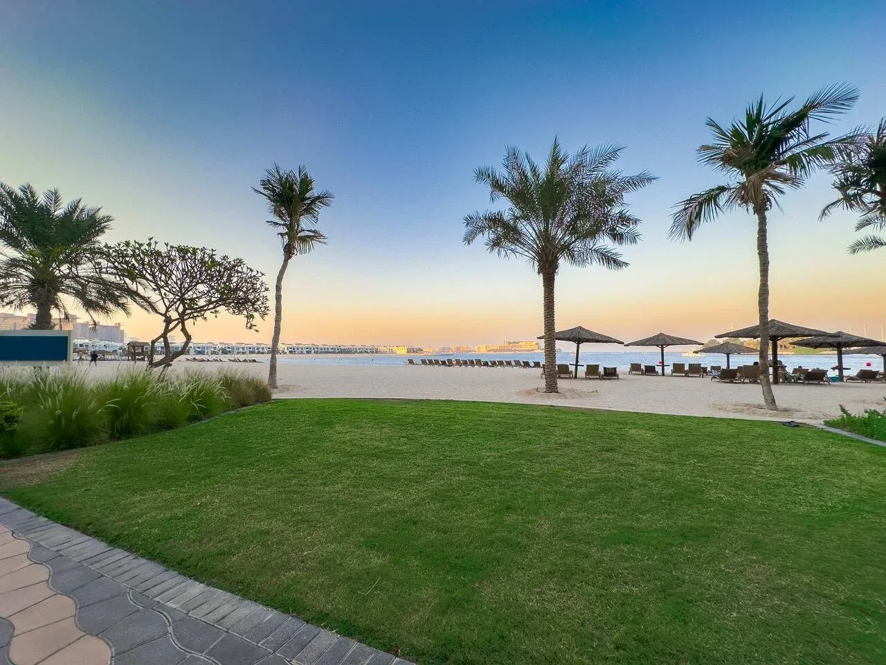 A landscaped waterfront lawn with palm trees and shaded seating at sunset.
