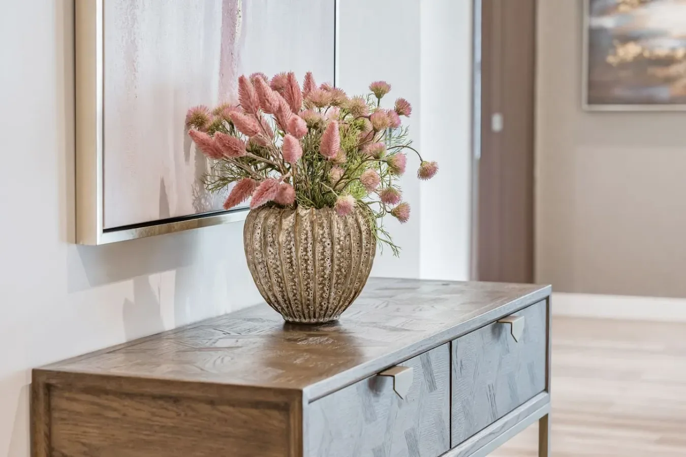 A decorative vase with flowers sits on a wooden console beneath framed artwork.