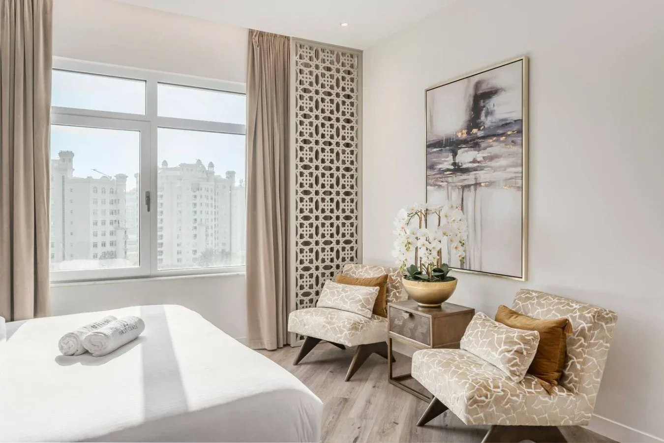 A bright bedroom has a large bed beside two patterned armchairs and a window.