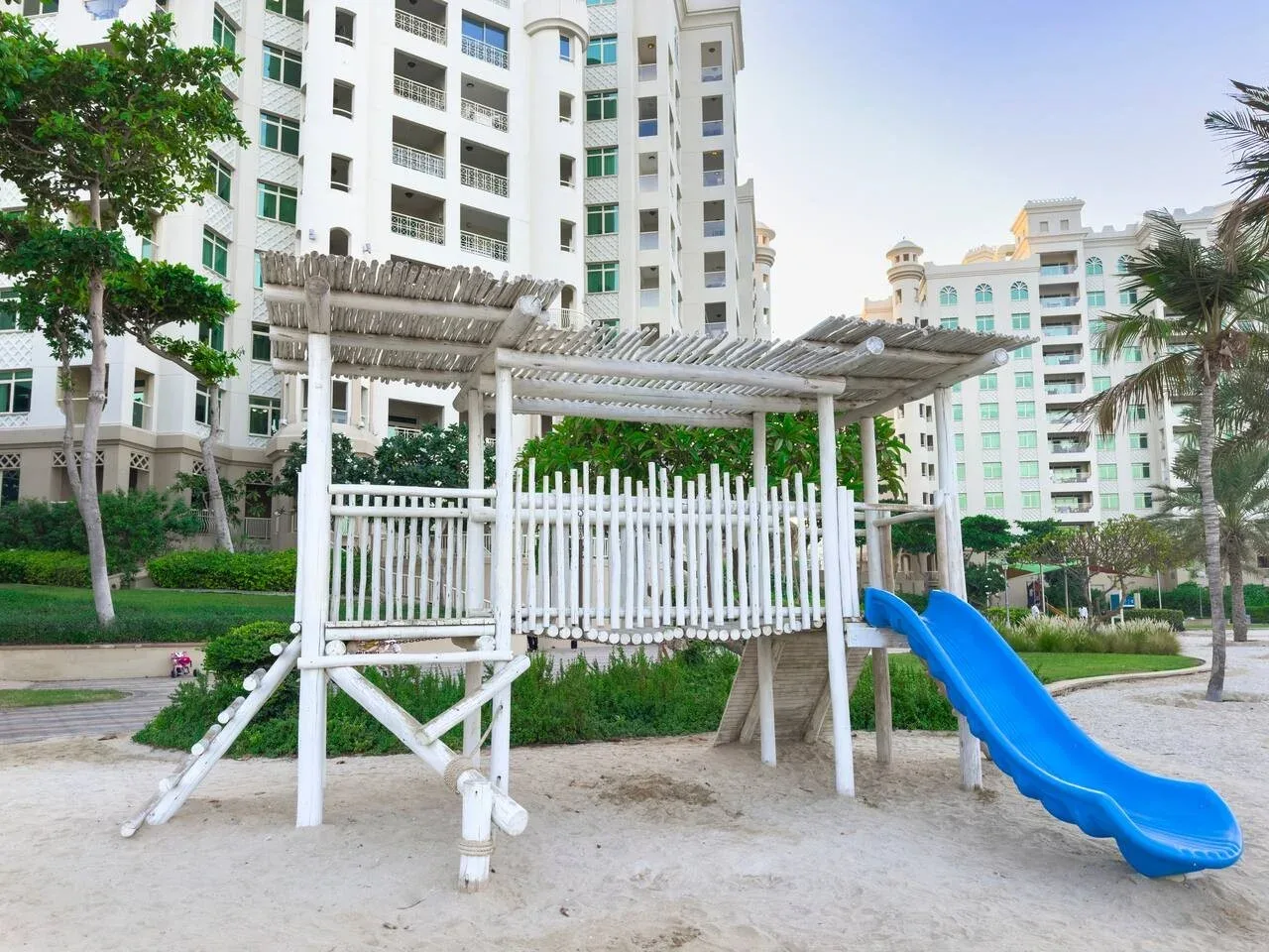 An outdoor playground with a blue slide beneath a white wooden climbing structure.