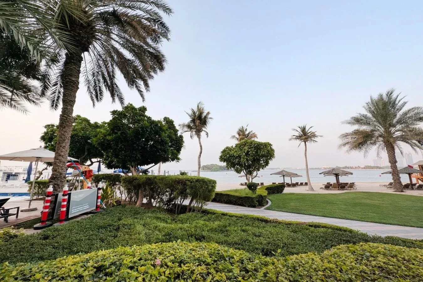 A landscaped waterfront area with palm trees, a path, and beach umbrellas.