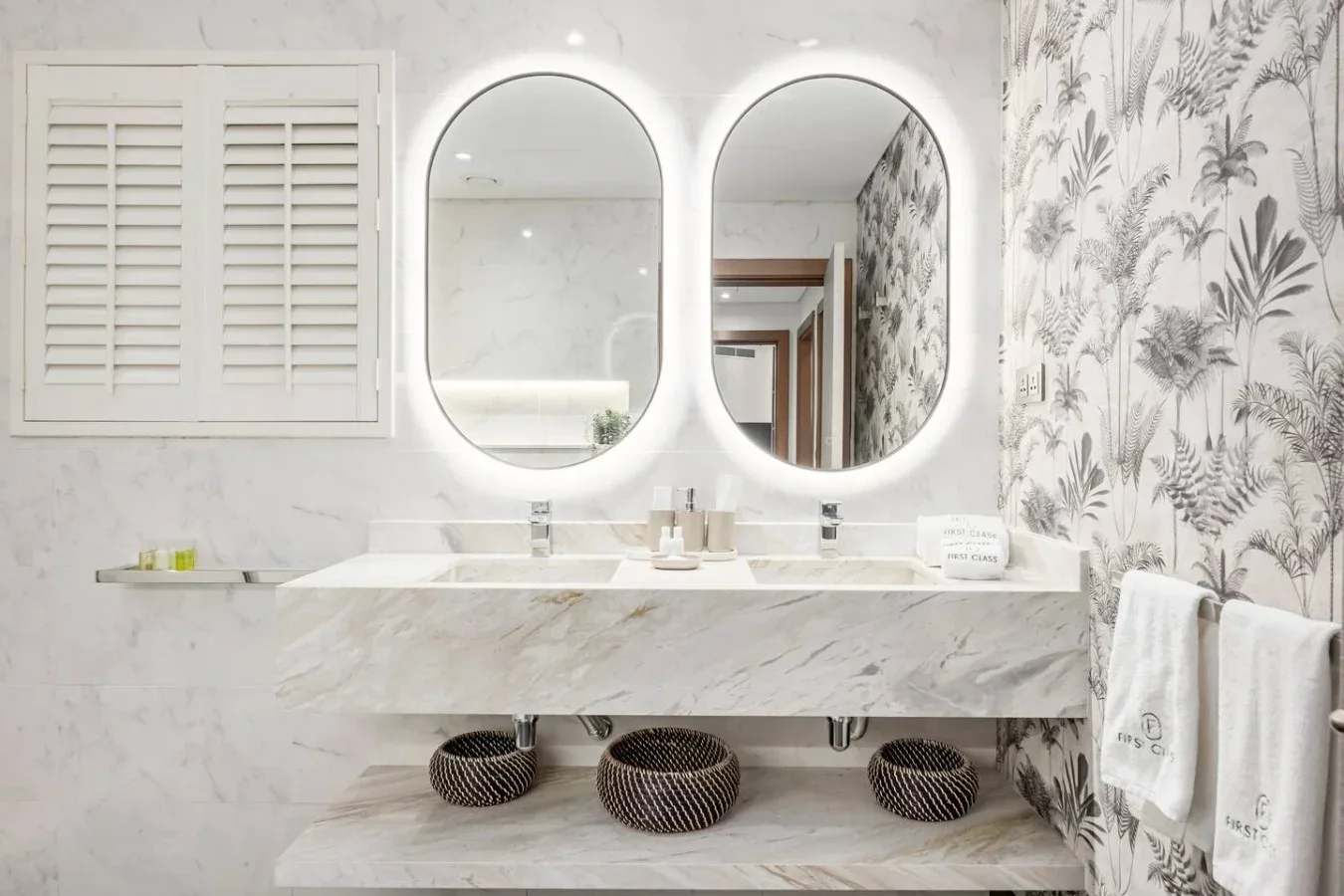 A bright bathroom vanity with two illuminated mirrors and patterned wallpaper.