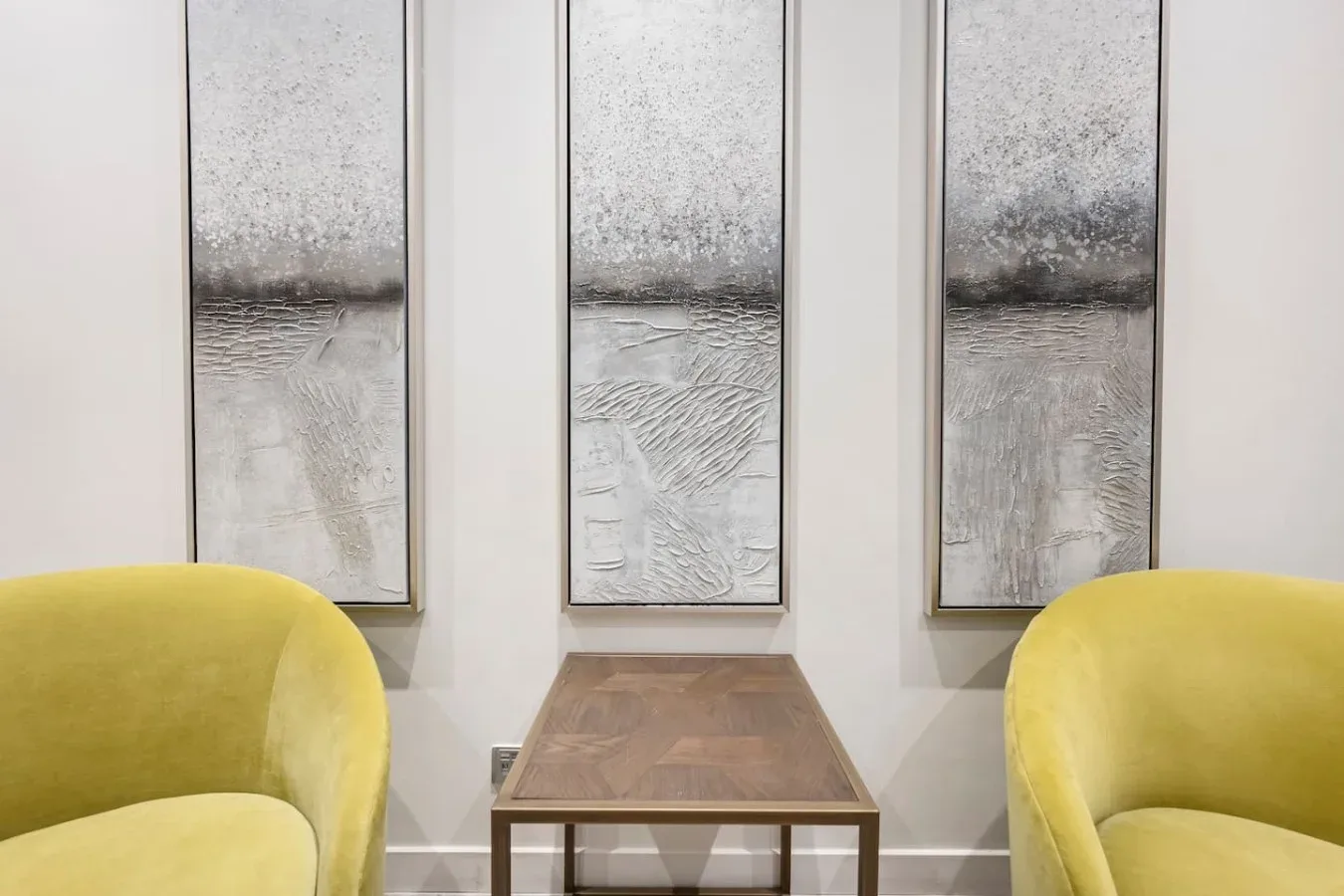 A seating area with yellow chairs and a small table beneath three abstract wall panels.