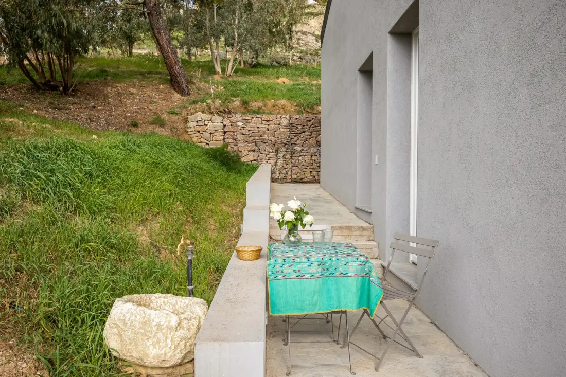 An outdoor patio beside a gray building, with a table set near a grassy slope.