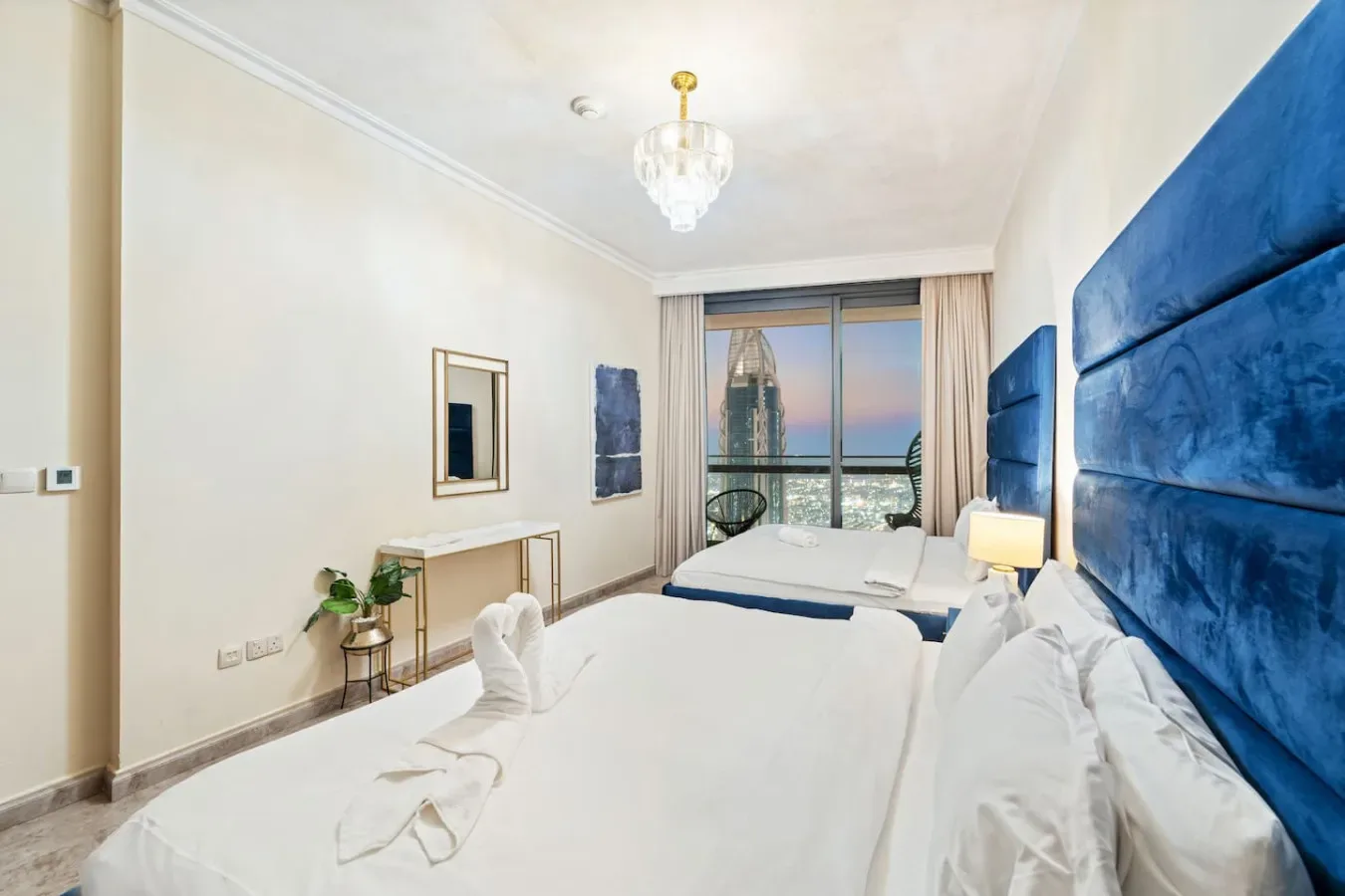 A bright bedroom with two beds, blue upholstered headboards, and a large window.
