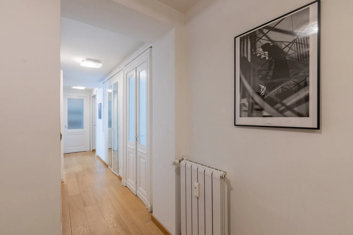 Un pasillo luminoso con puertas blancas, suelo de madera clara y un cuadro enmarcado.