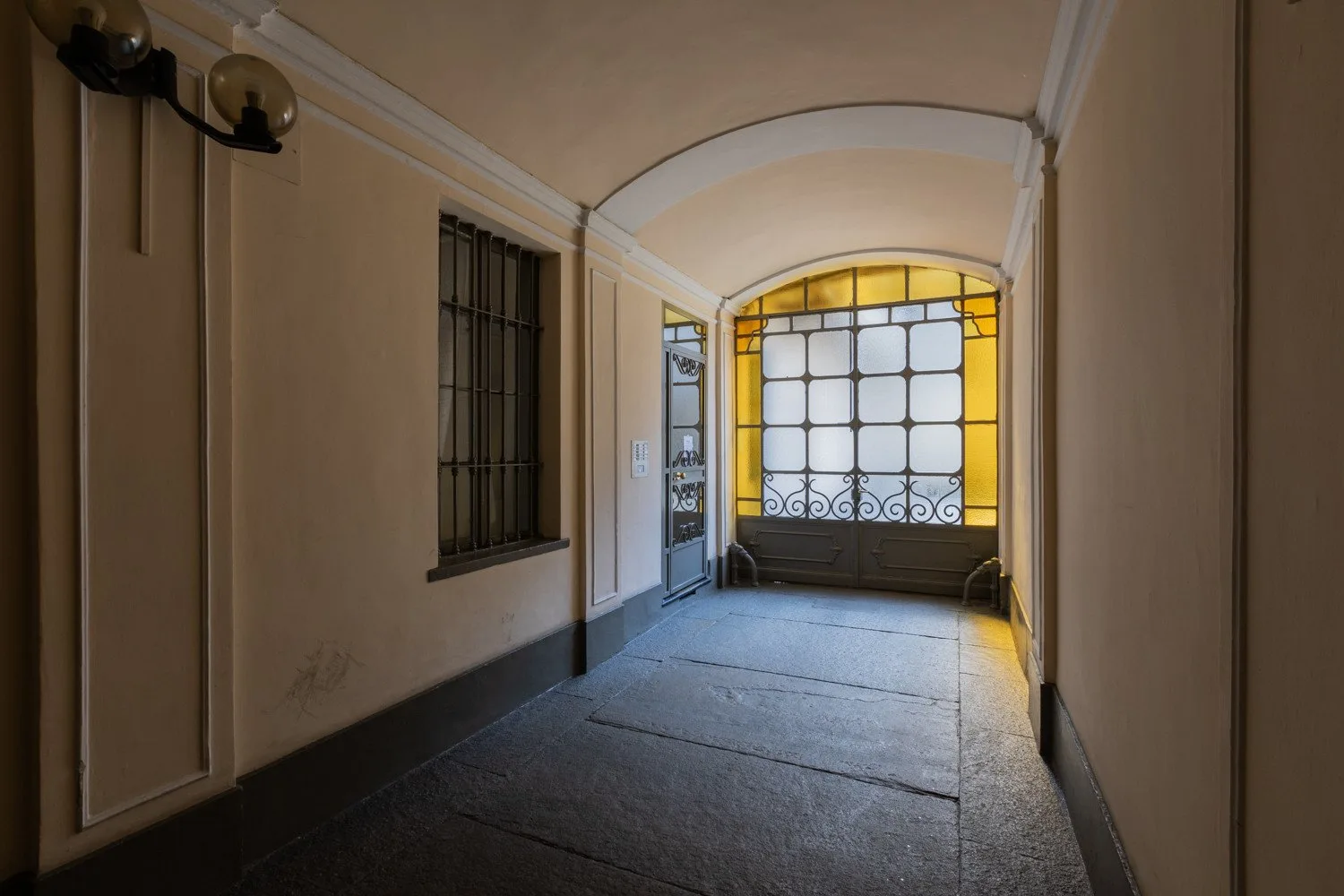 Un pasillo interior estrecho termina en una gran ventana con rejas metálicas oscuras.