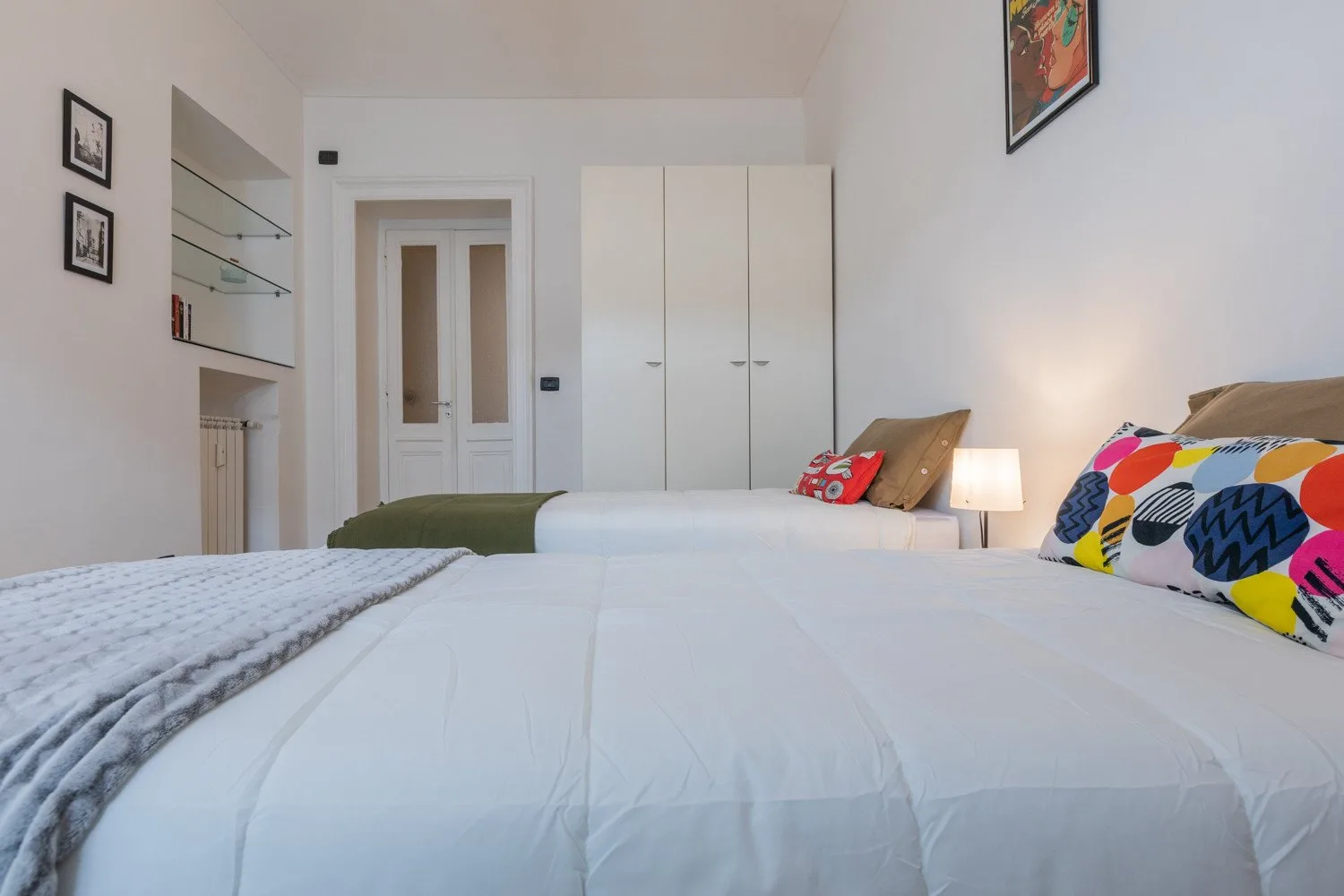 Un dormitorio con ropa de cama blanca, cojines coloridos y un armario.