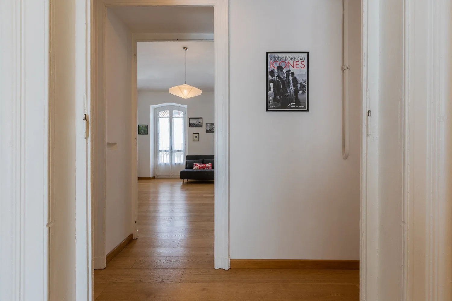 Un pasillo da a una habitación luminosa con suelo de madera y una lámpara colgante.