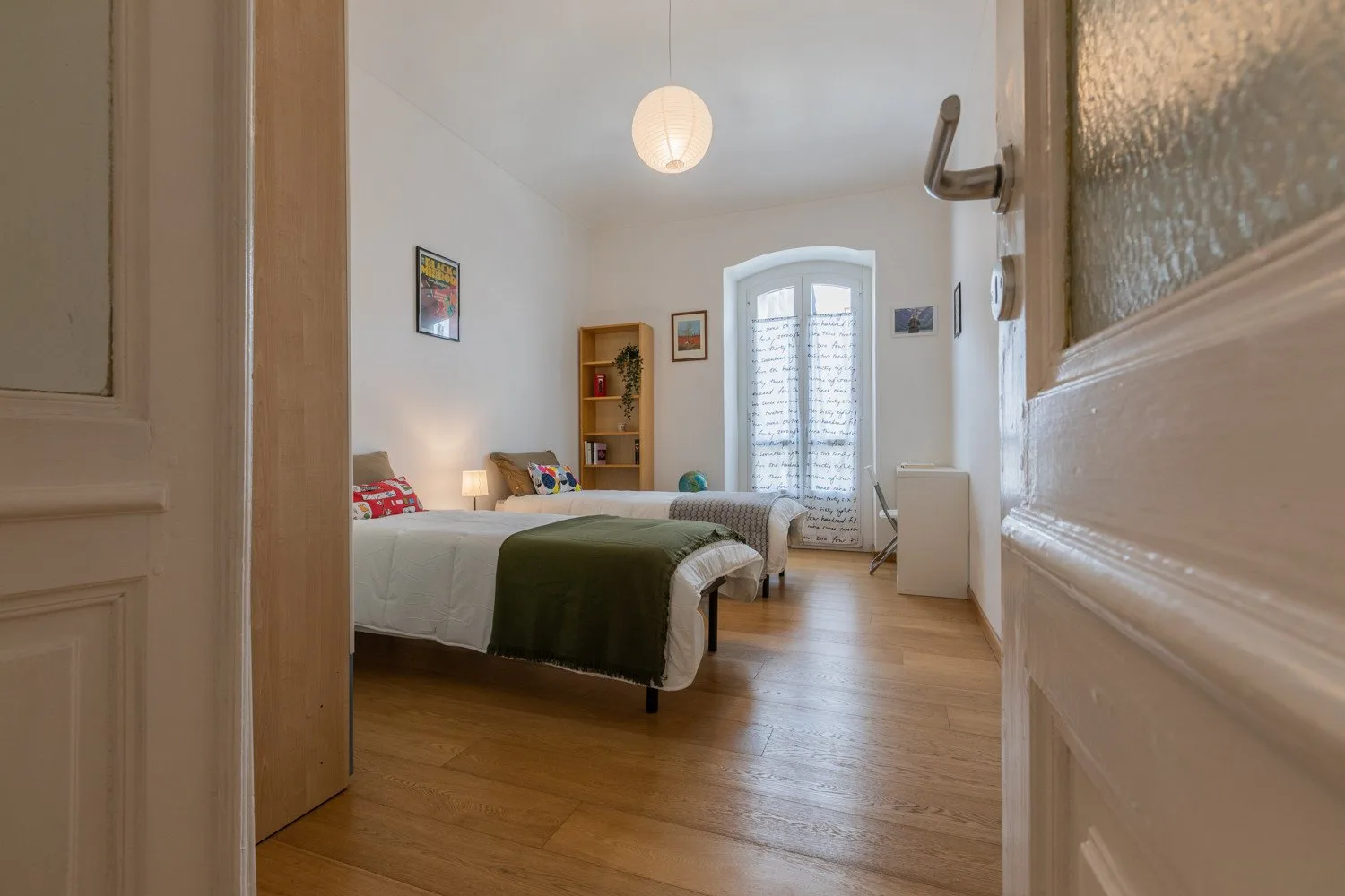 Un dormitorio con una cama, suelo de madera y una ventana alta.