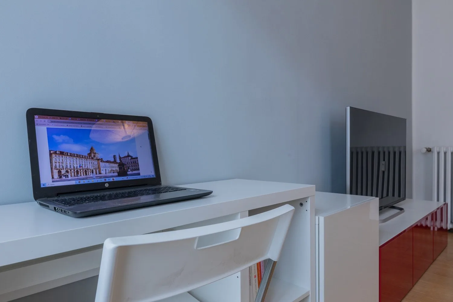 Un escritorio blanco con una computadora portátil y una silla está junto a un televisor.