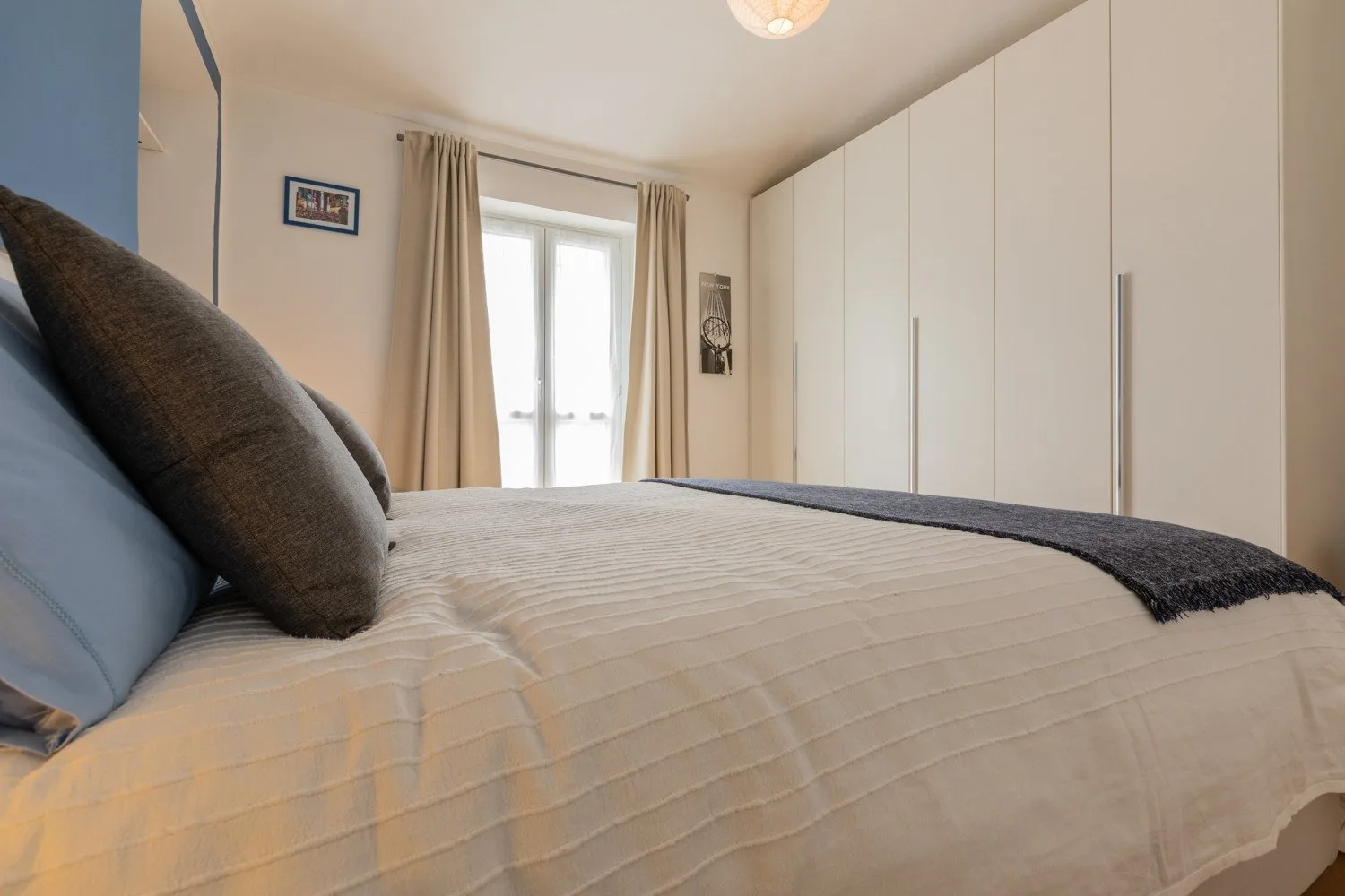 Un dormitorio con una cama bien tendida, armarios blancos altos y una ventana con cortinas.