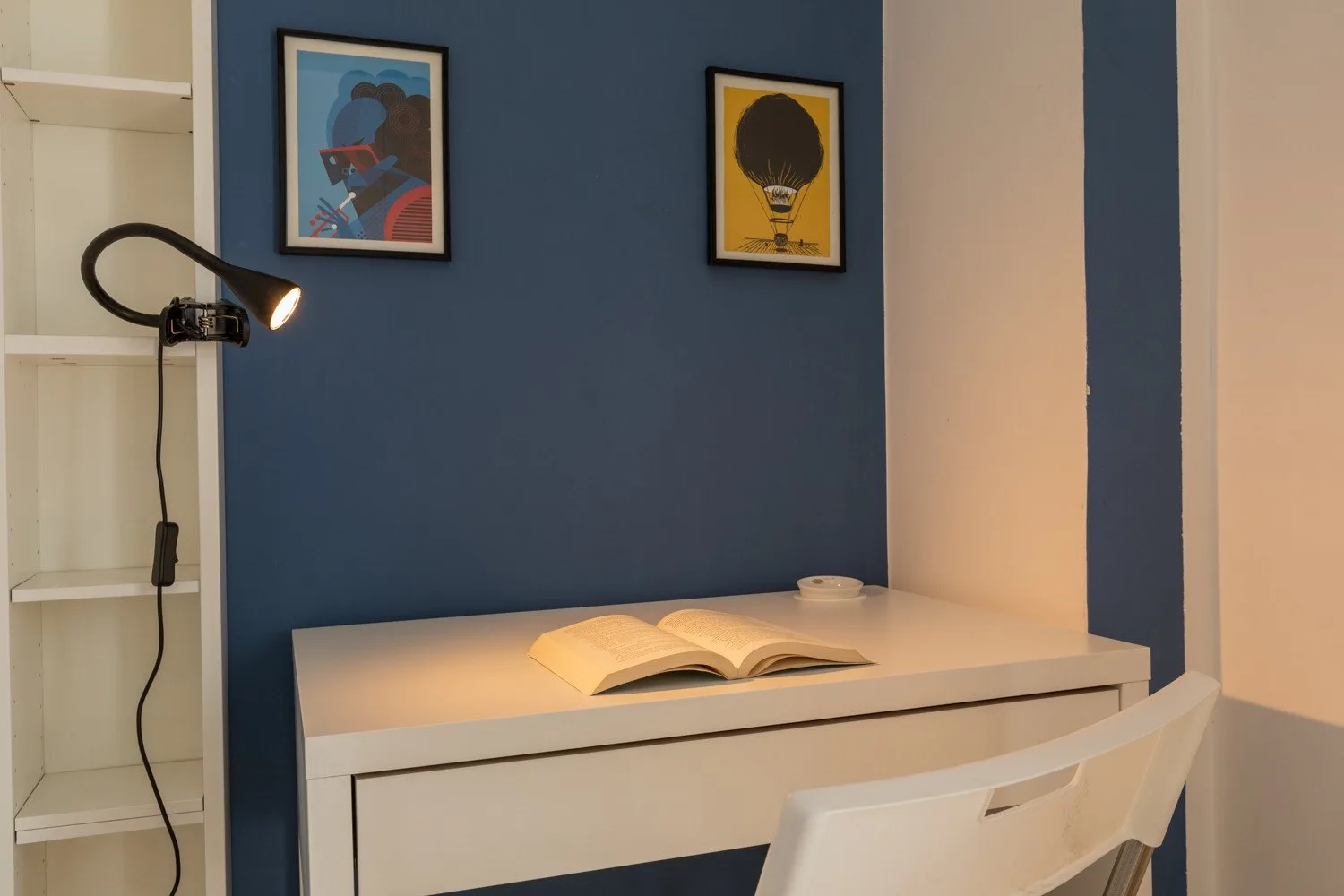 Un escritorio con un libro abierto está junto a una pared azul bajo dos láminas enmarcadas.