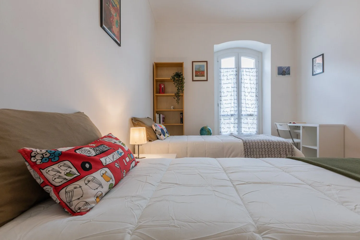 Un dormitorio con dos camas, una estantería de madera y una ventana alta con arco.