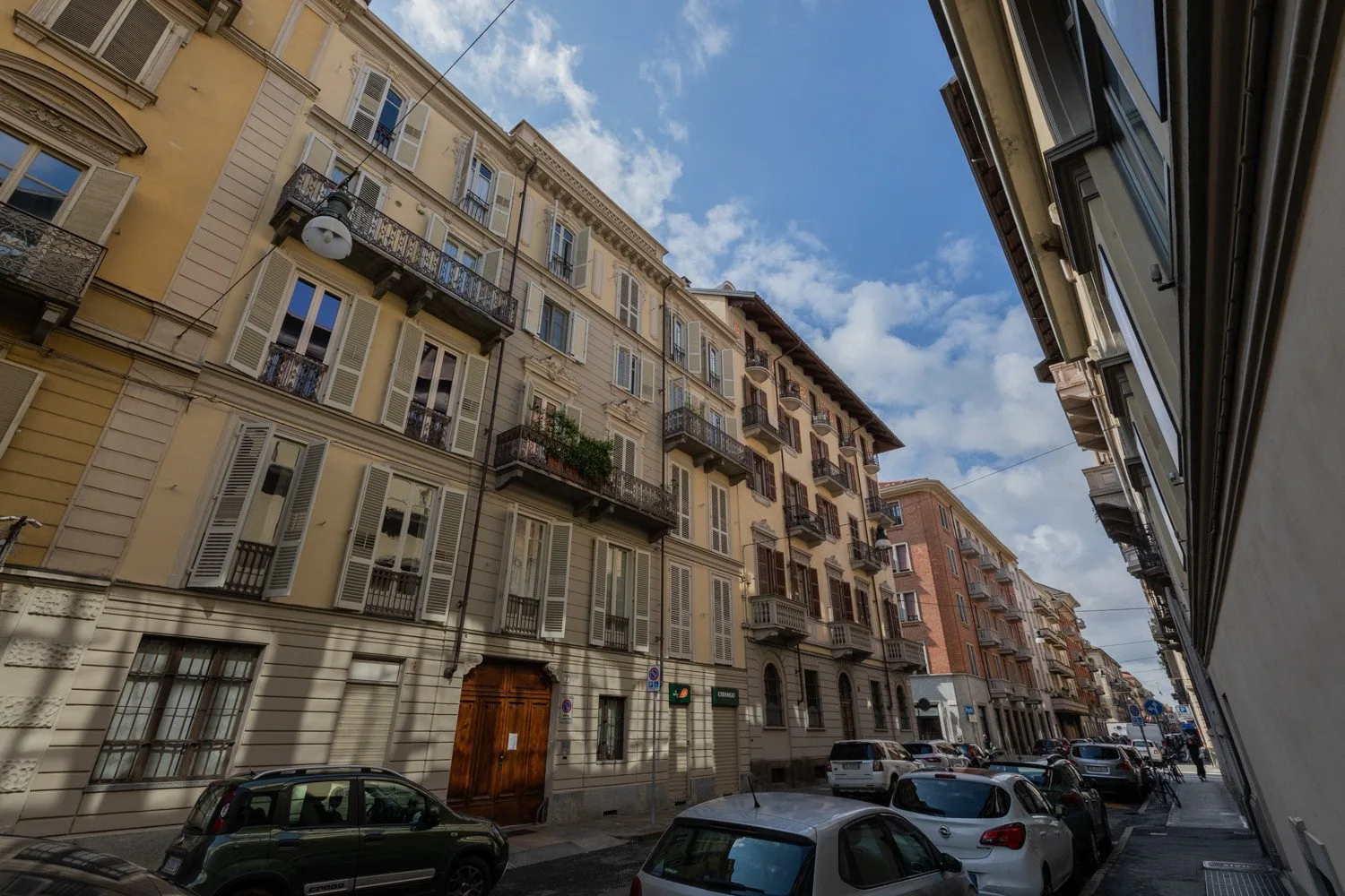 Una calle urbana con edificios de varias plantas y coches aparcados bajo un cielo azul.