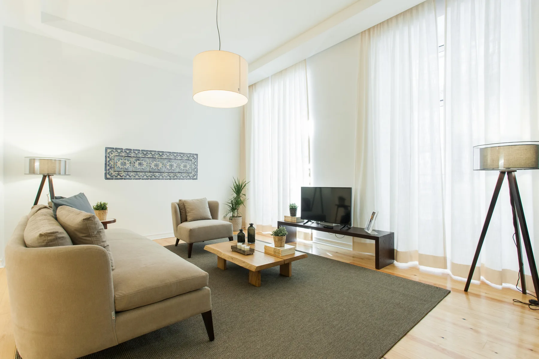 Una sala luminosa con asientos, una mesa de centro y un televisor junto a ventanas altas.