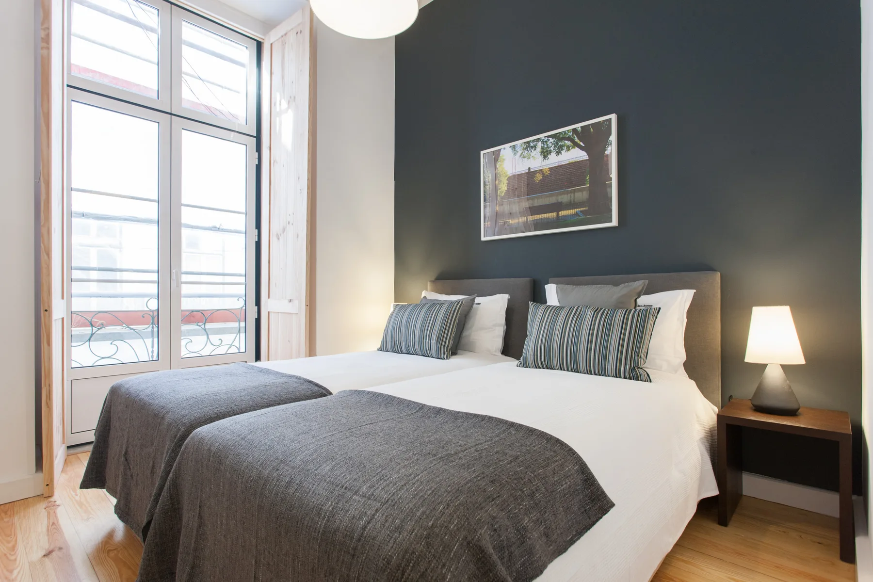 Dormitorio con una cama grande, ropa de cama gris y ventanas altas.