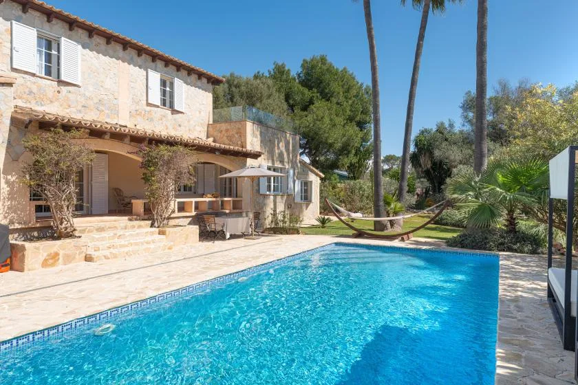 A swimming pool beside a stone house, with a hammock under palm trees.