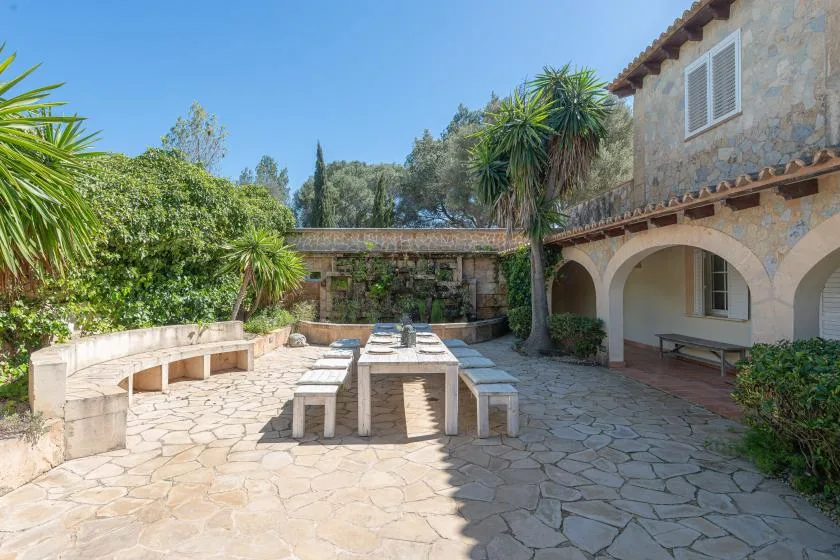 A stone courtyard with a long outdoor dining table surrounded by greenery.