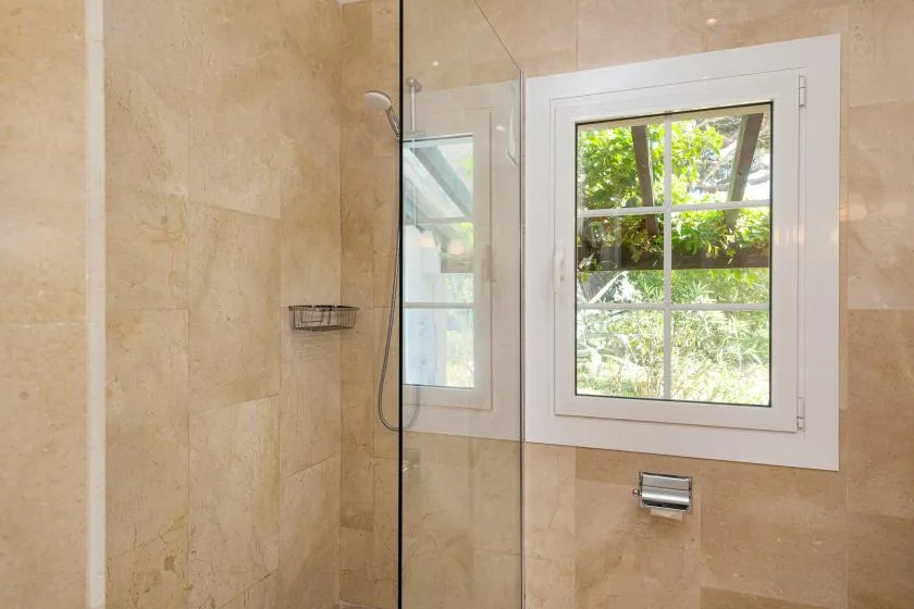 A tiled bathroom shower area beside a window overlooking green foliage.