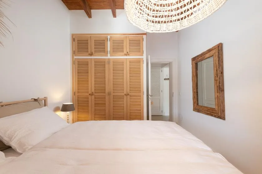 A bright bedroom with a bed, wooden shutters, and a woven pendant light.