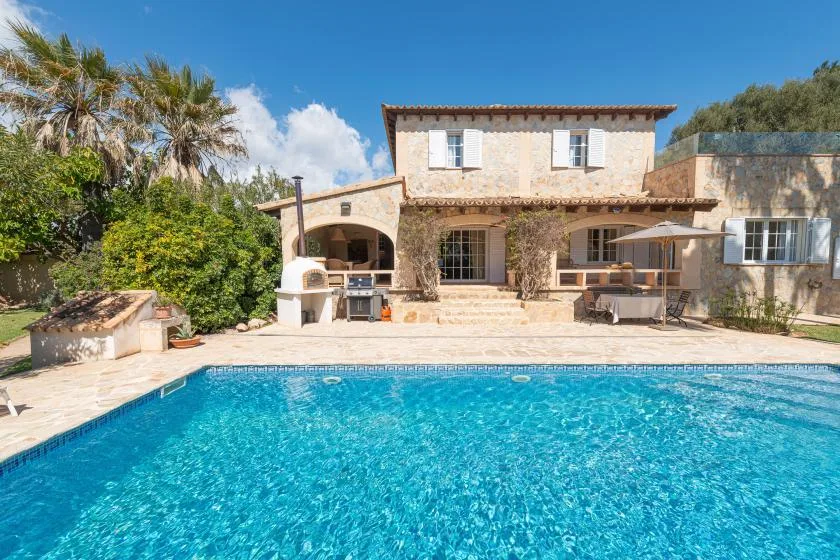 A swimming pool in front of a stone house with a shaded patio.