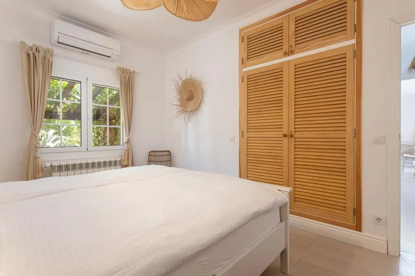 A bright bedroom with a large bed, curtained window, and wooden louvered closet.