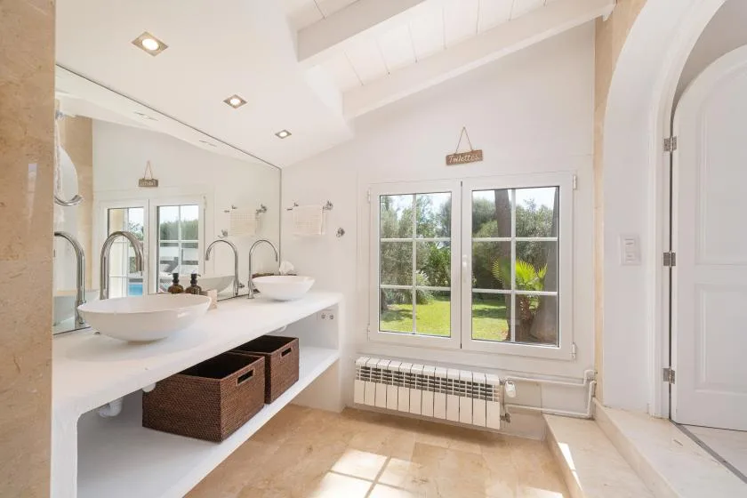 A bright bathroom with a double-sink vanity and windows overlooking greenery.
