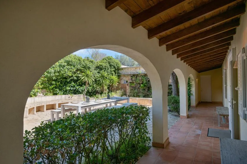 A covered outdoor walkway with arched openings overlooks a leafy garden.
