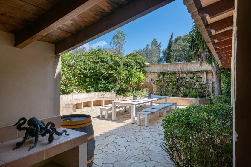 A paved outdoor patio with a long table and benches, surrounded by greenery.