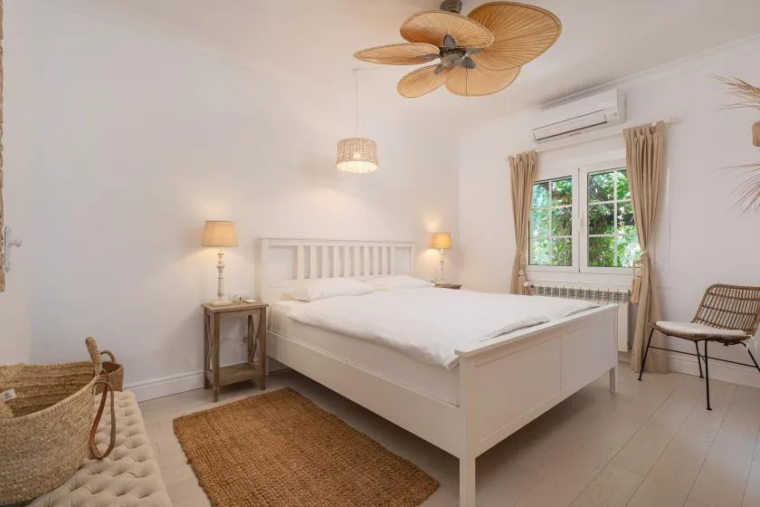 A bright bedroom with a white bed, bedside lamps, and a window overlooking greenery.