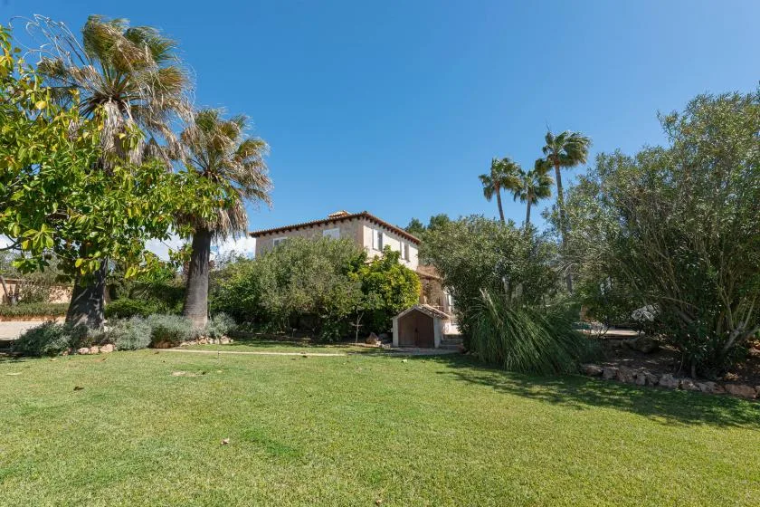 A house sits beyond a broad grassy lawn, with trees and shrubs around it.