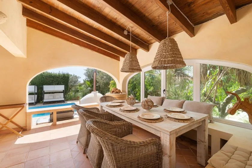 A dining area with a long table and woven seating beside large arched windows.