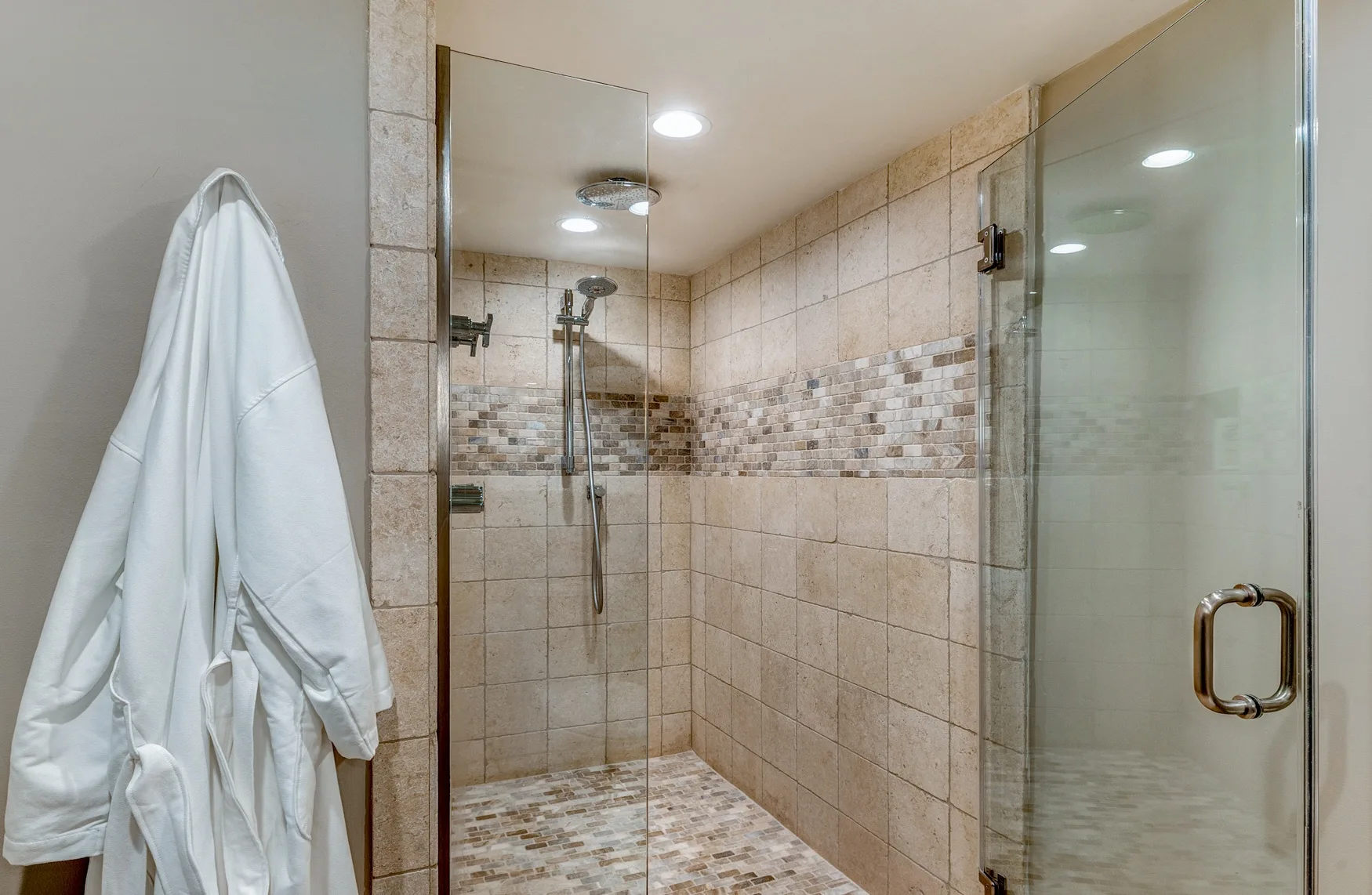 A tiled bathroom with a glass shower and a white robe hanging beside it.