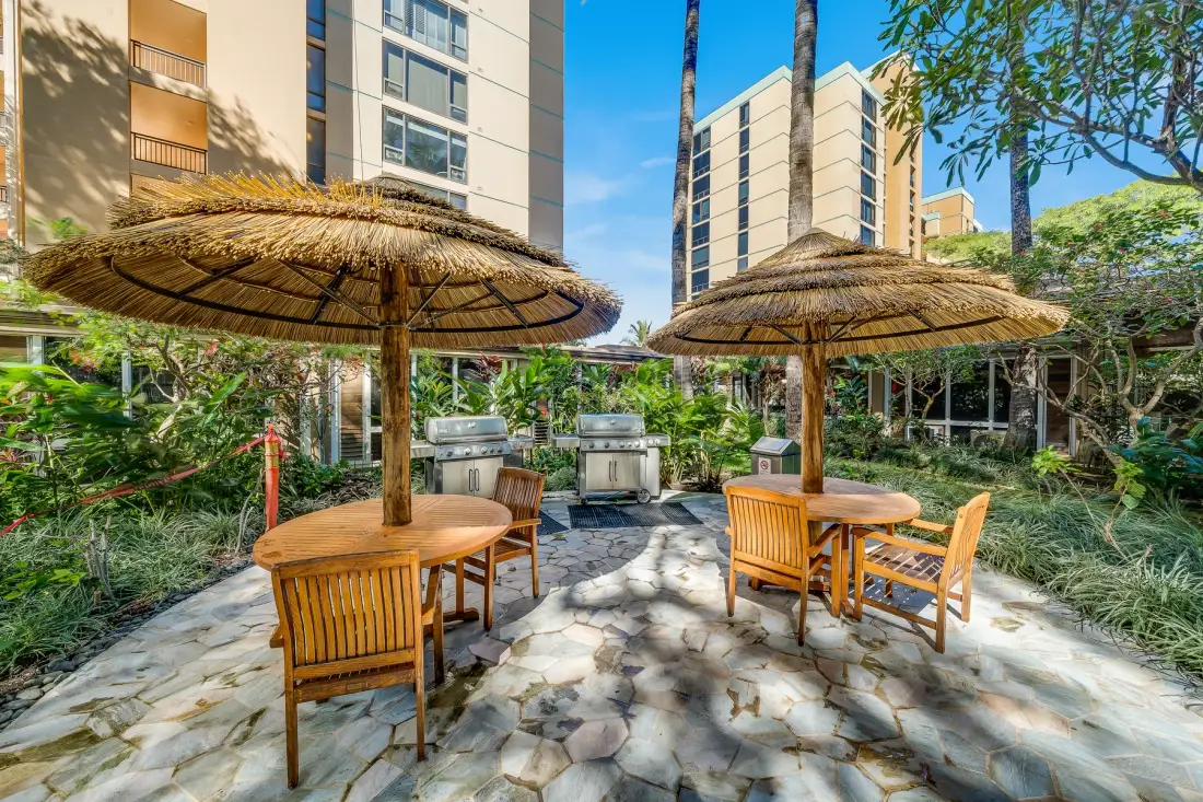 An outdoor patio with wooden seating beneath thatched parasols, surrounded by greenery.