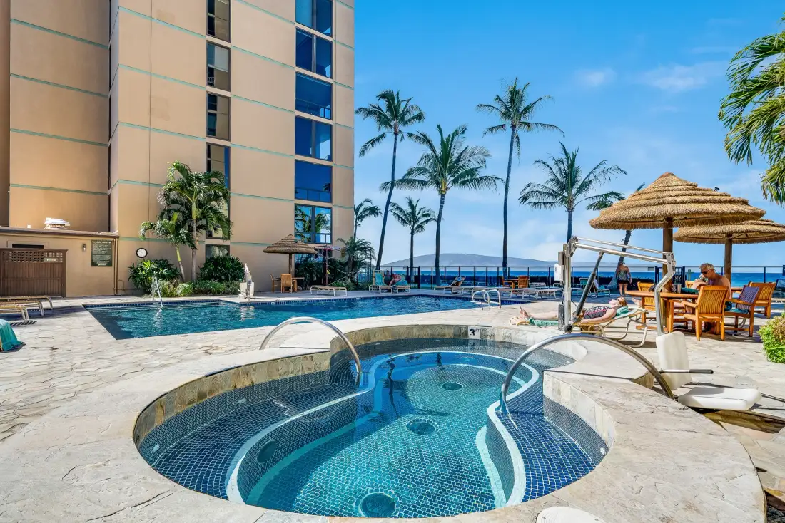 An outdoor pool and circular spa sit beside a palm-lined beachfront building.
